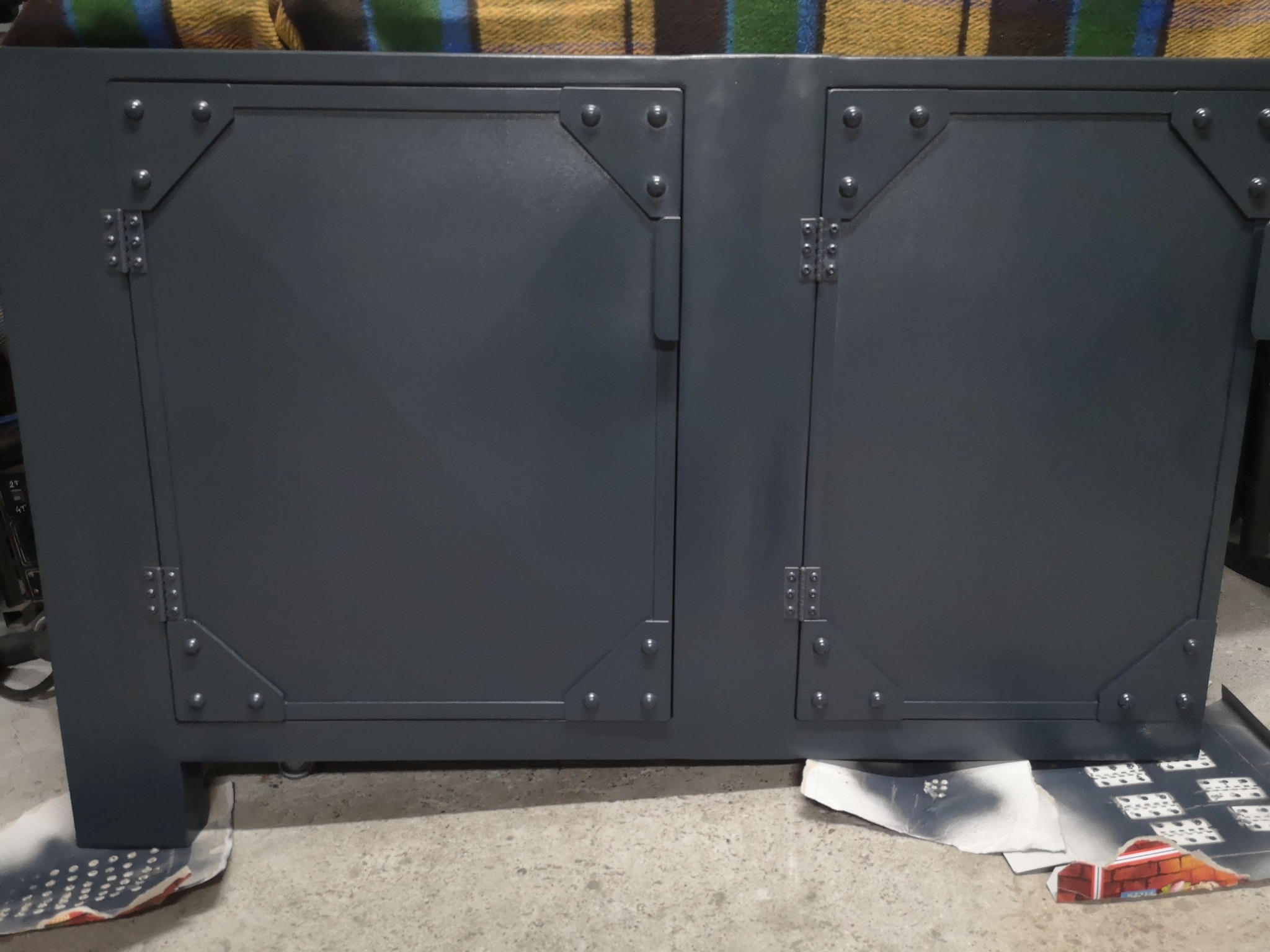 Metalowa szafka z dwoma drzwiczkami, wzmocniona nitowanymi narożnikami, pomalowana na szaro, stoi na kartonach w warsztacie.