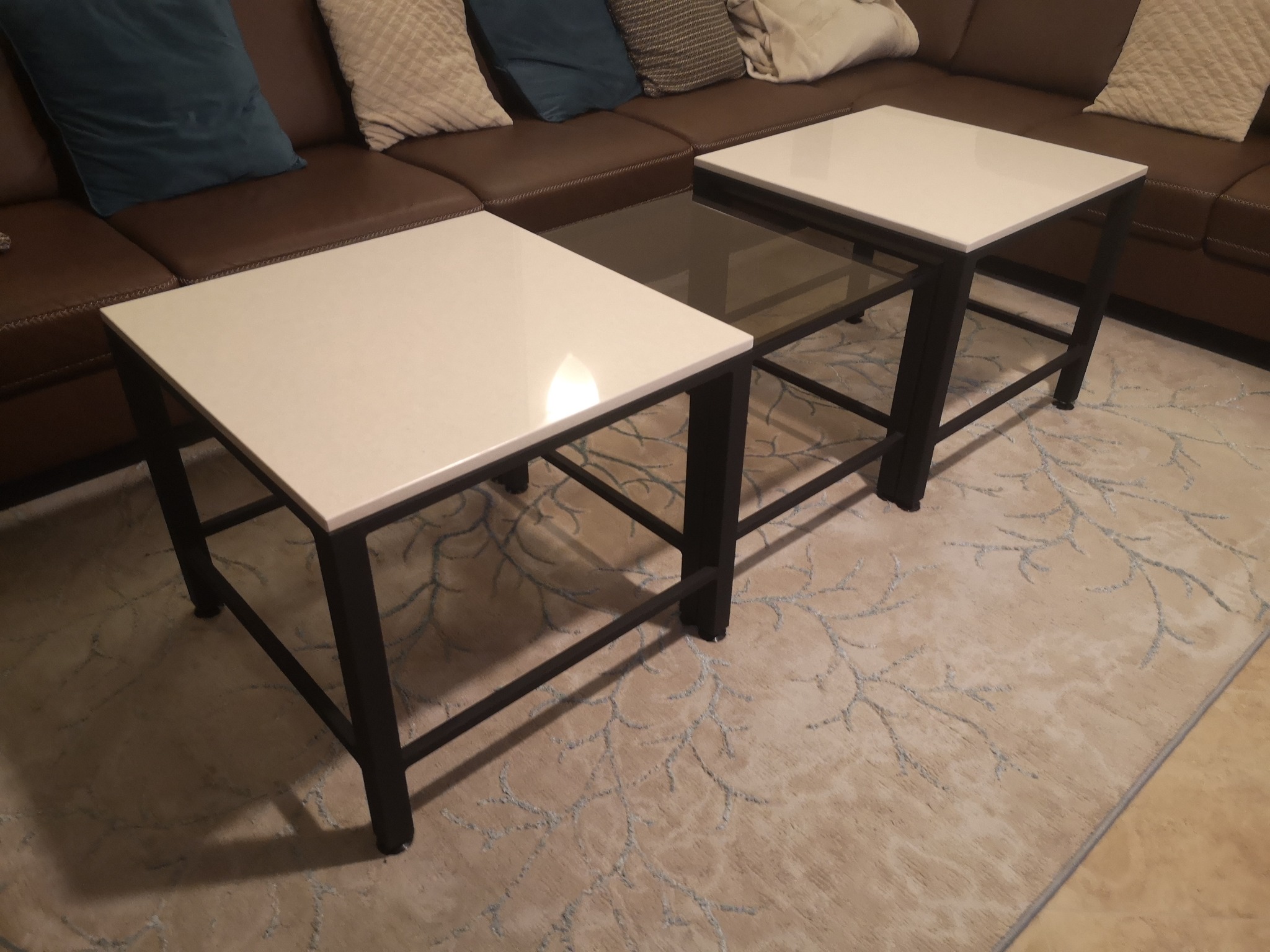 Dwa kwadratowe stoliki kawowe z białymi blatami i czarnymi metalowymi ramami, jeden z dodatkową szklaną półką, ustawione na wzorzystym dywanie przed brązową skórzaną sofą z poduszkami.
