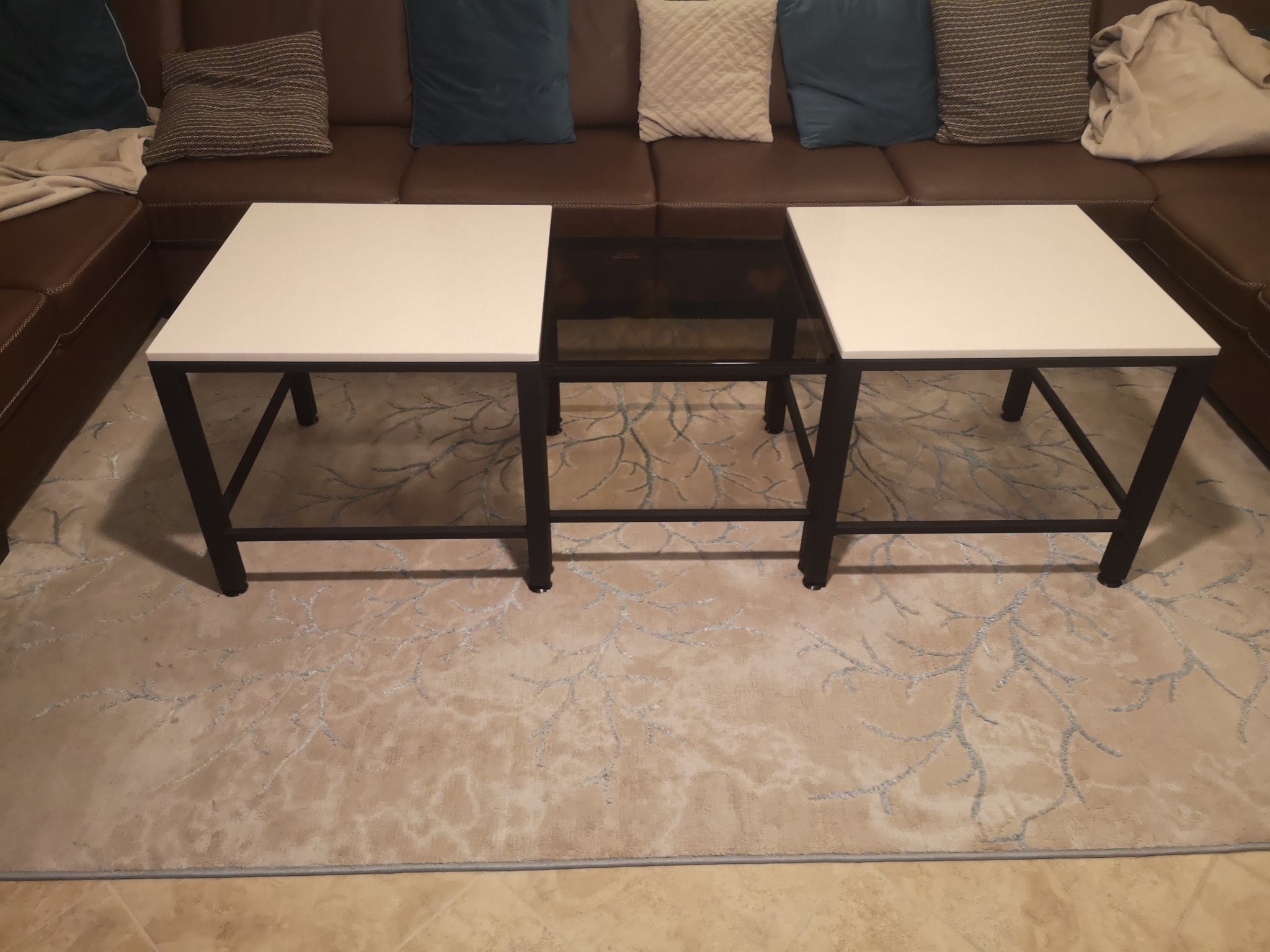 Nowoczesny zestaw trzech stolików kawowych z metalowymi nogami i blatami z białego laminatu oraz ciemnego szkła, ustawiony na beżowym dywanie w salonie z brązową sofą narożną i dekoracyjnymi...