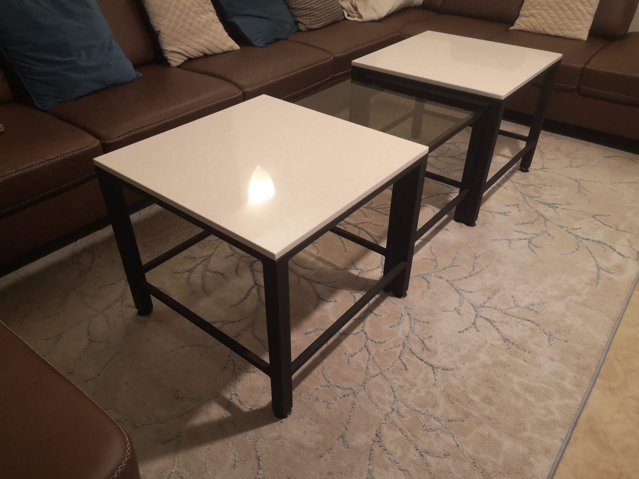 Dwa nowoczesne stoliki kawowe z białymi blatami i czarnymi metalowymi ramami, ustawione na wzorzystym dywanie w salonie z brązową sofą w tle.