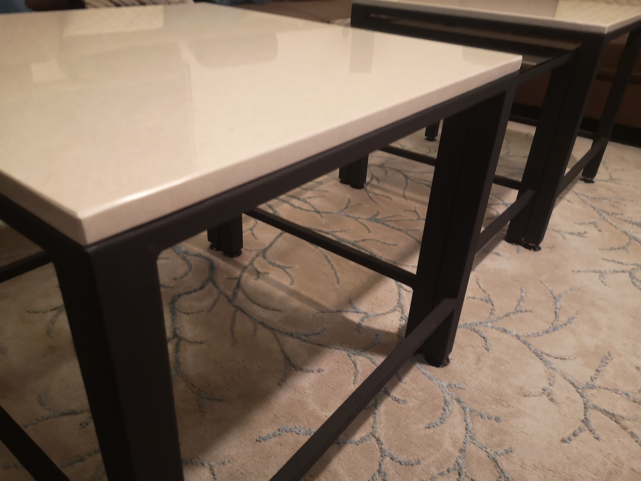 Dwa stoliki kawowe z metalową ramą i jasnym blatem, ustawione na beżowym dywanie z niebieskim wzorem roślinnym.