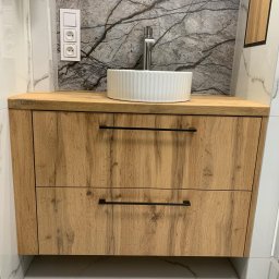 Meble na wymiar - Łazienkowa szafka pod umywalkę z dwoma szufladami o drewnianym wykończeniu, biała umywalka nablatowa z chromowaną baterią, marmurowe płytki na ścianie i podłodze, widoczne gniazdka elektryczne.