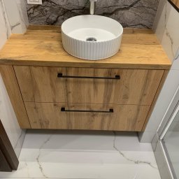 Meble na wymiar - Biała, okrągła umywalka nablatowa na drewnianej szafce z dwoma szufladami z czarnymi uchwytami w łazience w Białymstoku, marmurowe płytki na ścianie i podłodze.