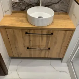 Biała, okrągła umywalka nablatowa na drewnianej szafce z dwoma szufladami z czarnymi uchwytami w łazience w Białymstoku, marmurowe płytki na ścianie i podłodze.