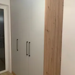 Wysoka szafa z szarymi frontami i drewnianym bokiem, z czarnymi, pionowymi uchwytami, widoczna z boku przy oknie.