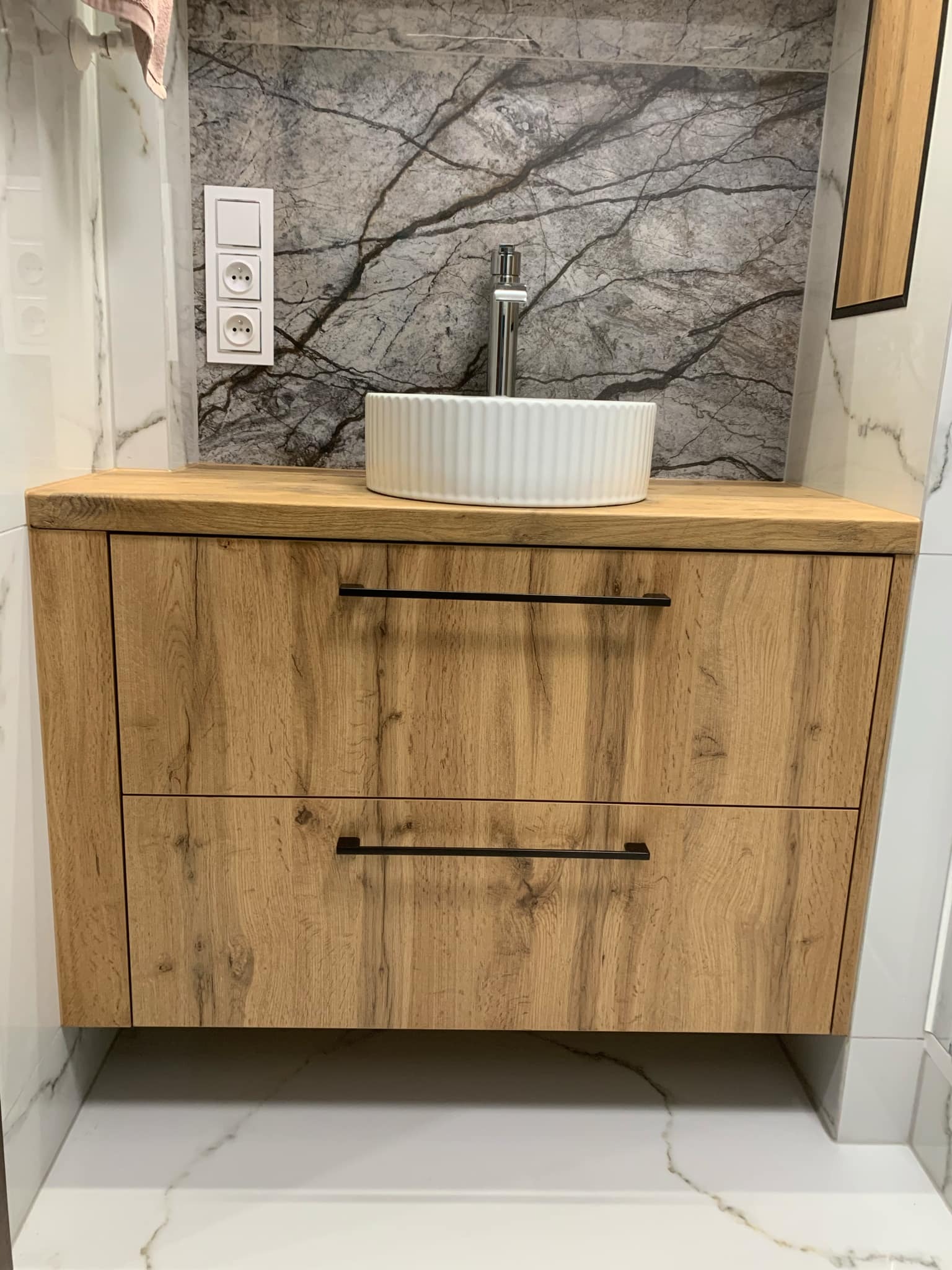 Łazienkowa szafka pod umywalkę z dwoma szufladami o drewnianym wykończeniu, biała umywalka nablatowa z chromowaną baterią, marmurowe płytki na ścianie i podłodze, widoczne gniazdka elektryczne.