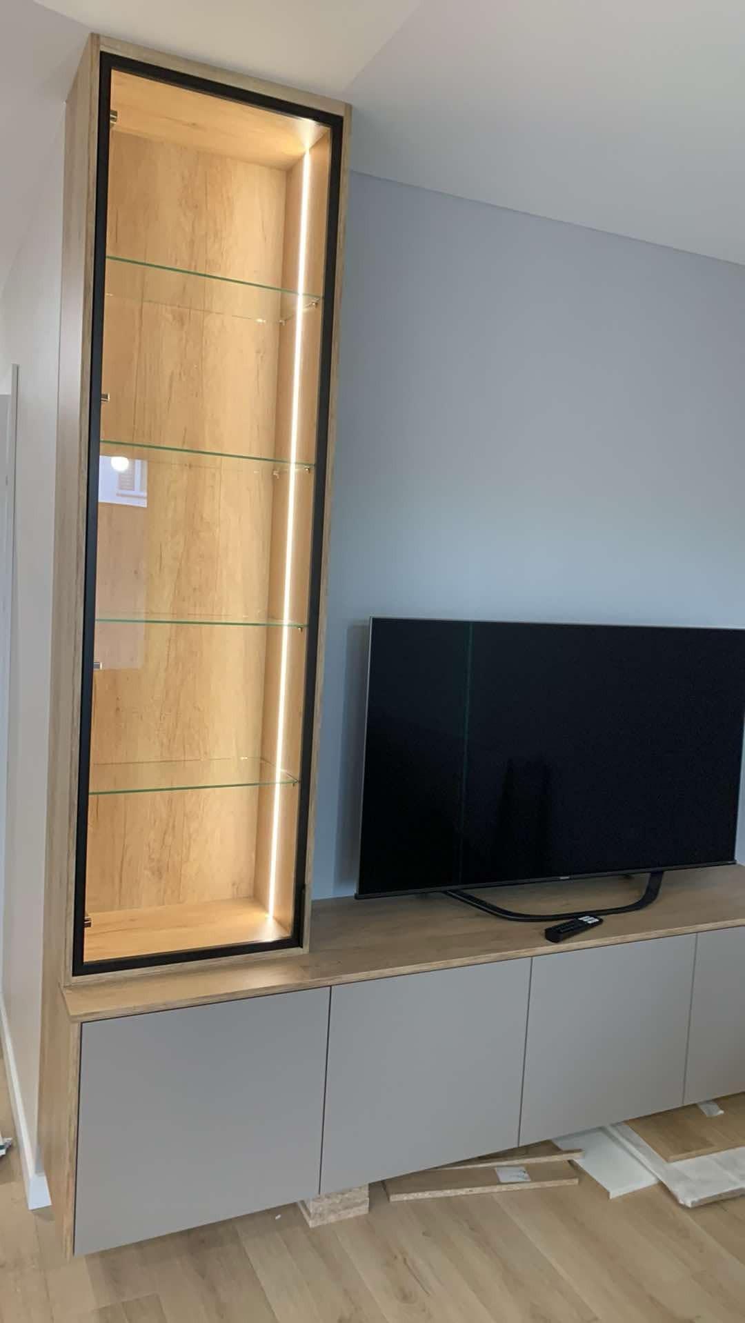 Nowoczesna szafka RTV z podświetlaną witryną z drewnopodobnym wykończeniem i szklanymi półkami, na tle błękitnej ściany i telewizora.