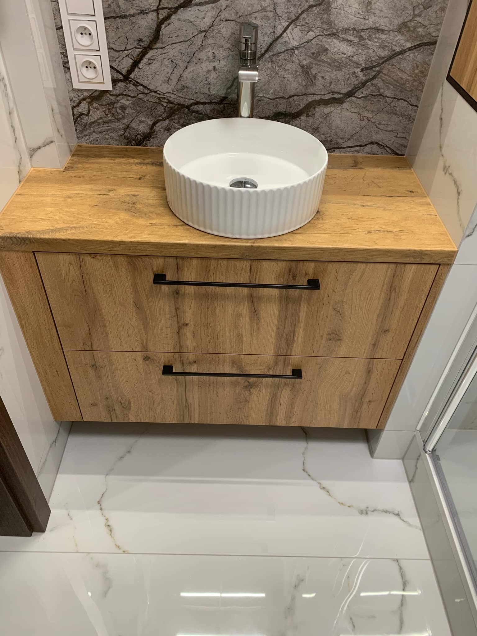 Biała, okrągła umywalka nablatowa na drewnianej szafce z dwoma szufladami z czarnymi uchwytami w łazience w Białymstoku, marmurowe płytki na ścianie i podłodze.