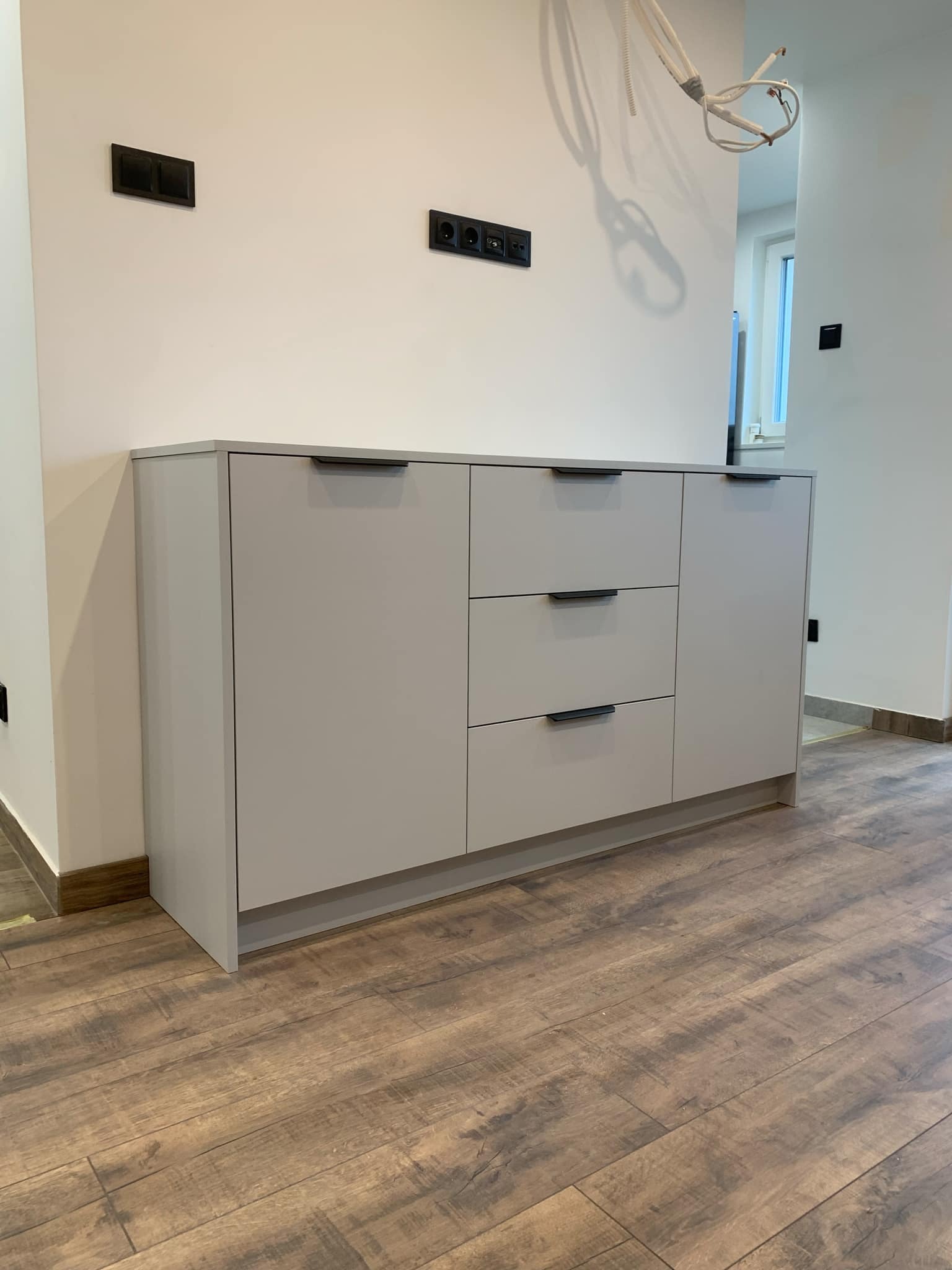 Jasnoszara szafka z trzema szufladami i dwojgiem drzwiczek, z czarnymi uchwytami, na tle białej ściany z czarnymi gniazdkami elektrycznymi, na podłodze panele imitujące drewno, widoczne kable...