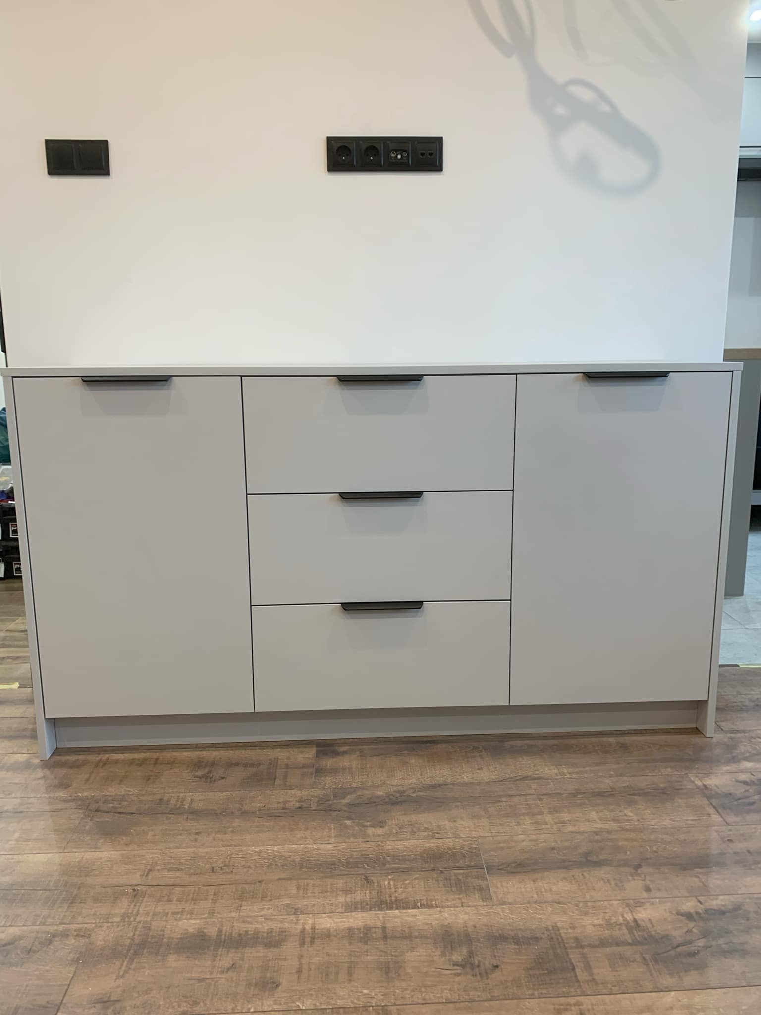 Jasnoszara komoda z trzema szufladami i dwojgiem drzwi z czarnymi uchwytami, na tle białej ściany z gniazdkami i drewnianej podłogi.