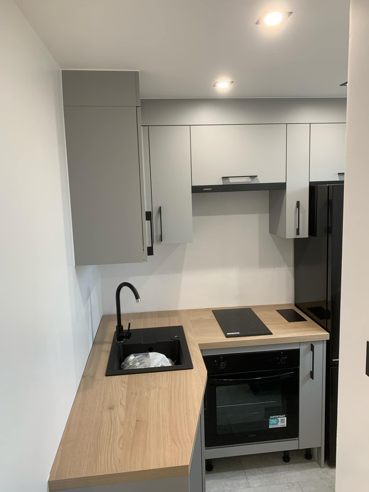 Nowoczesna, szara kuchnia z drewnianym blatem, czarną armaturą i płytą indukcyjną. W zlewie widoczne opakowania. Ujęcie z perspektywy stojącej osoby.