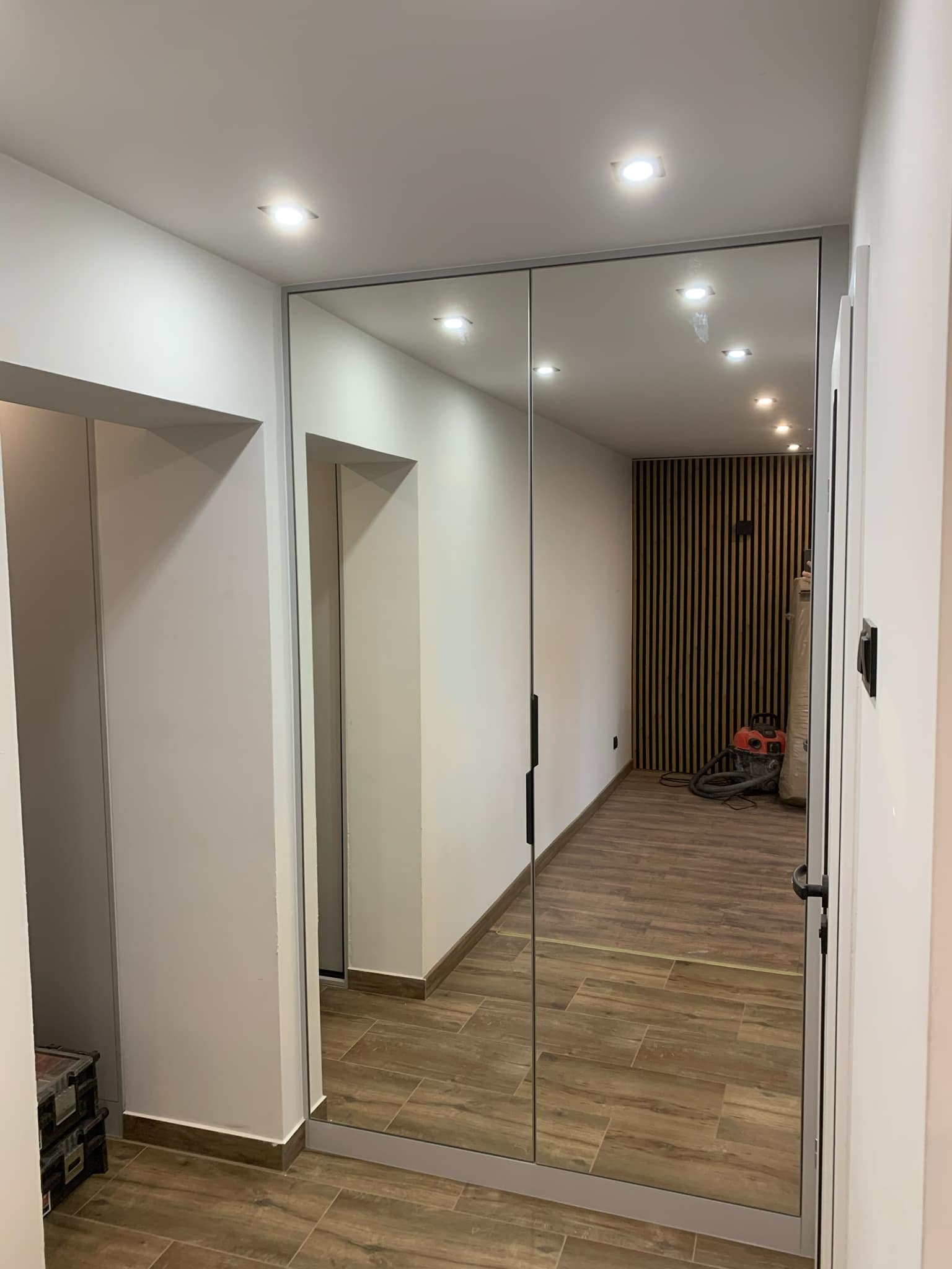 Nowoczesna szafa wnękowa z lustrzanymi drzwiami przesuwnymi, odbijająca wnętrze z drewnianą boazerią i odkurzaczem, widoczna skrzynka narzędziowa w rogu.