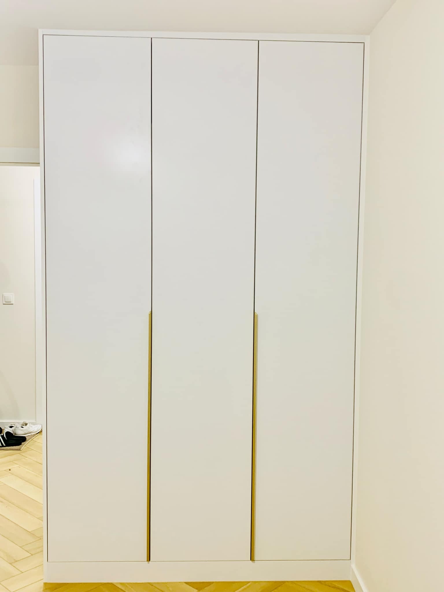 Trzydrzwiowa, biała szafa wnękowa z minimalistycznymi, złotymi uchwytami, wkomponowana w jasne wnętrze z parkietem w jodełkę.
