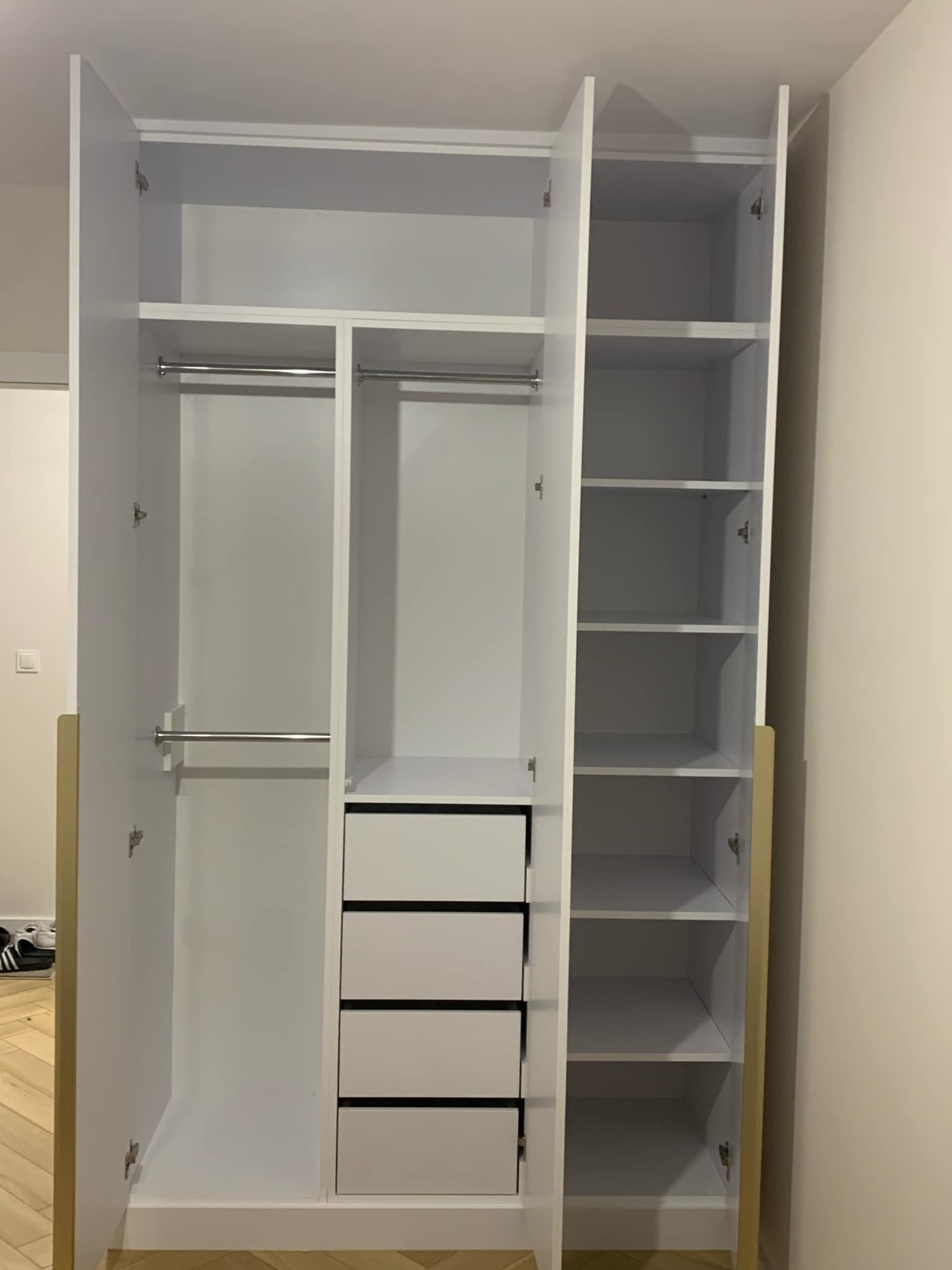 Wnętrze białej szafy z otwartymi drzwiami, widoczne półki, drążki na ubrania i cztery szuflady z czarnymi uchwytami, umieszczone w dolnej części szafy.