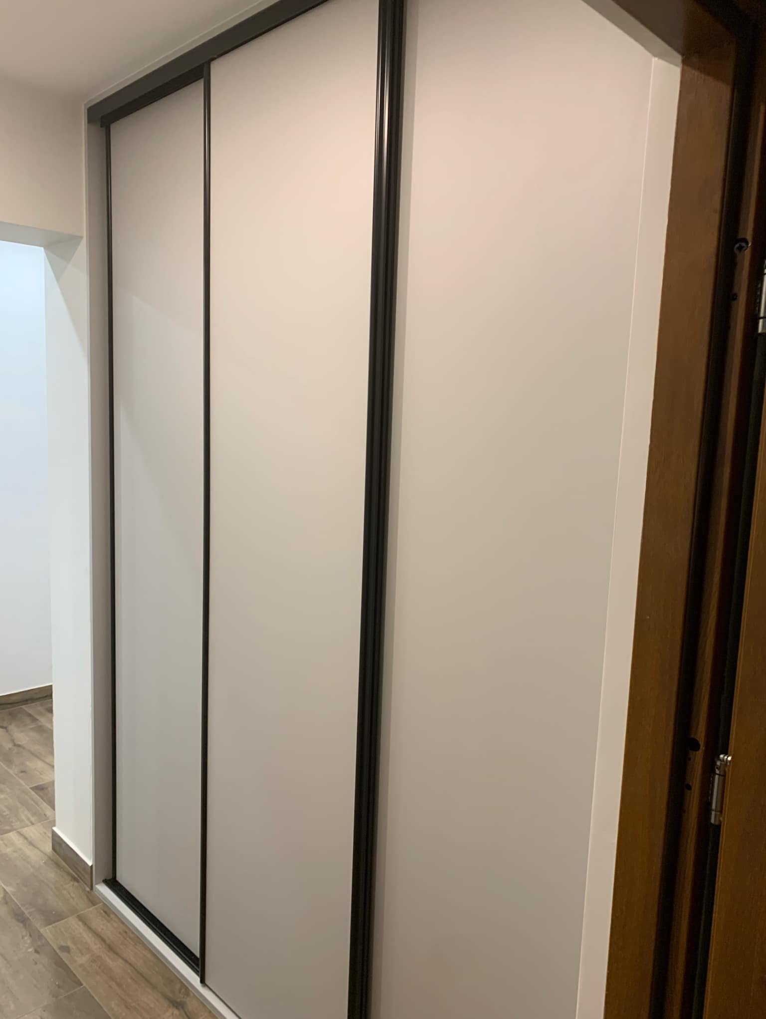 Trzydrzwiowa szafa przesuwna z białymi frontami i czarnymi obramowaniami, widoczna w korytarzu z drewnopodobną podłogą i brązowymi drzwiami po prawej stronie.