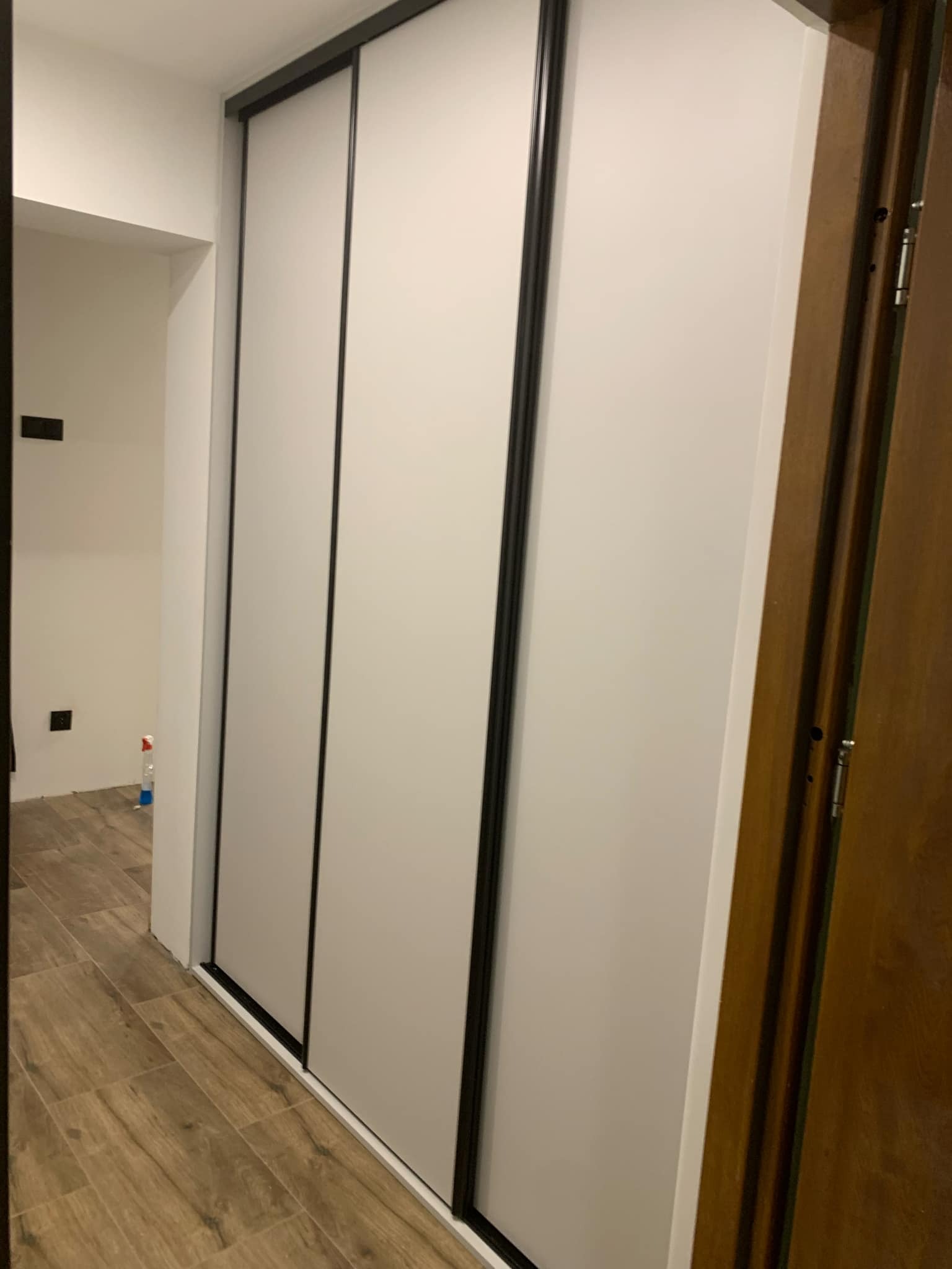 Biała szafa przesuwna z czarnymi obramowaniami drzwi, widoczna z boku w korytarzu z drewnopodobną podłogą i brązowymi drzwiami po prawej stronie.
