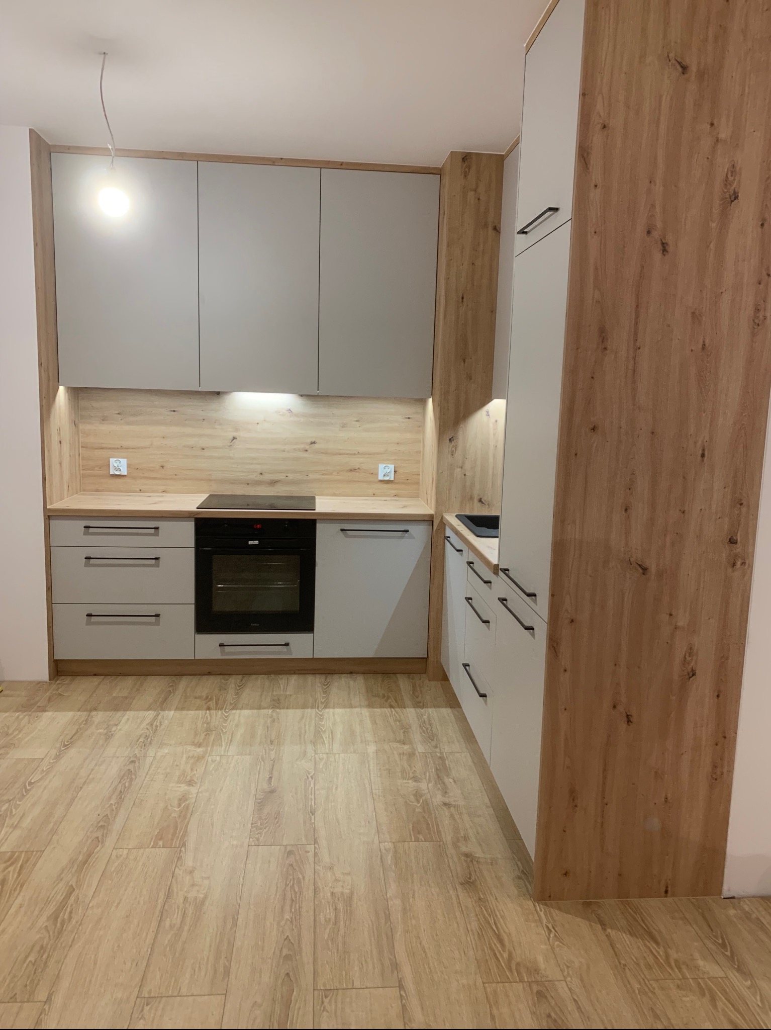 Nowoczesna kuchnia z szarymi frontami szafek i drewnianym wykończeniem, widoczna płyta indukcyjna i piekarnik, oświetlenie podszafkowe, podłoga imitująca drewno.