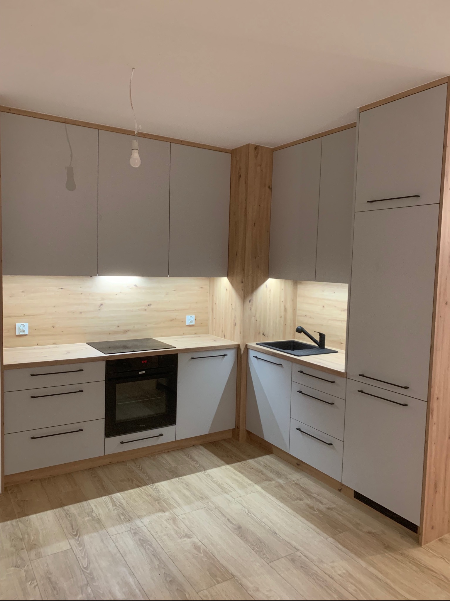 Jasnoszara kuchnia na wymiar z drewnianymi akcentami w Białymstoku, z czarnym zlewem i minimalistycznymi uchwytami, oświetlona listwą LED pod szafkami.