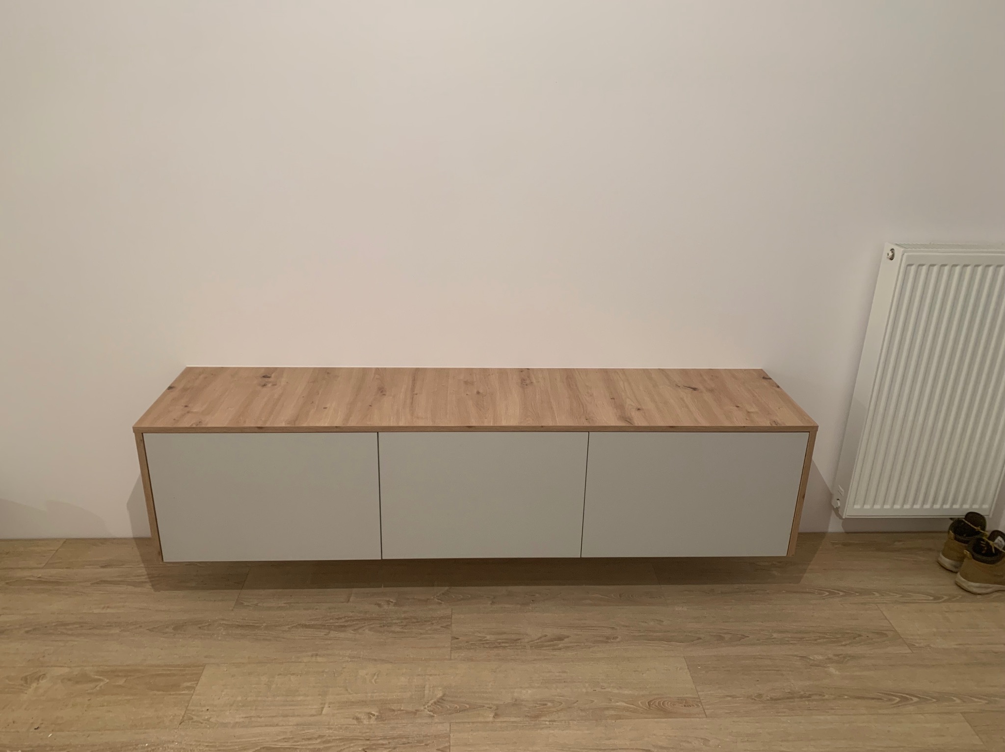 Wisząca szafka RTV z trzema szarymi frontami i blatem w kolorze jasnego drewna, zamontowana na białej ścianie, z widocznym grzejnikiem i butami w tle.