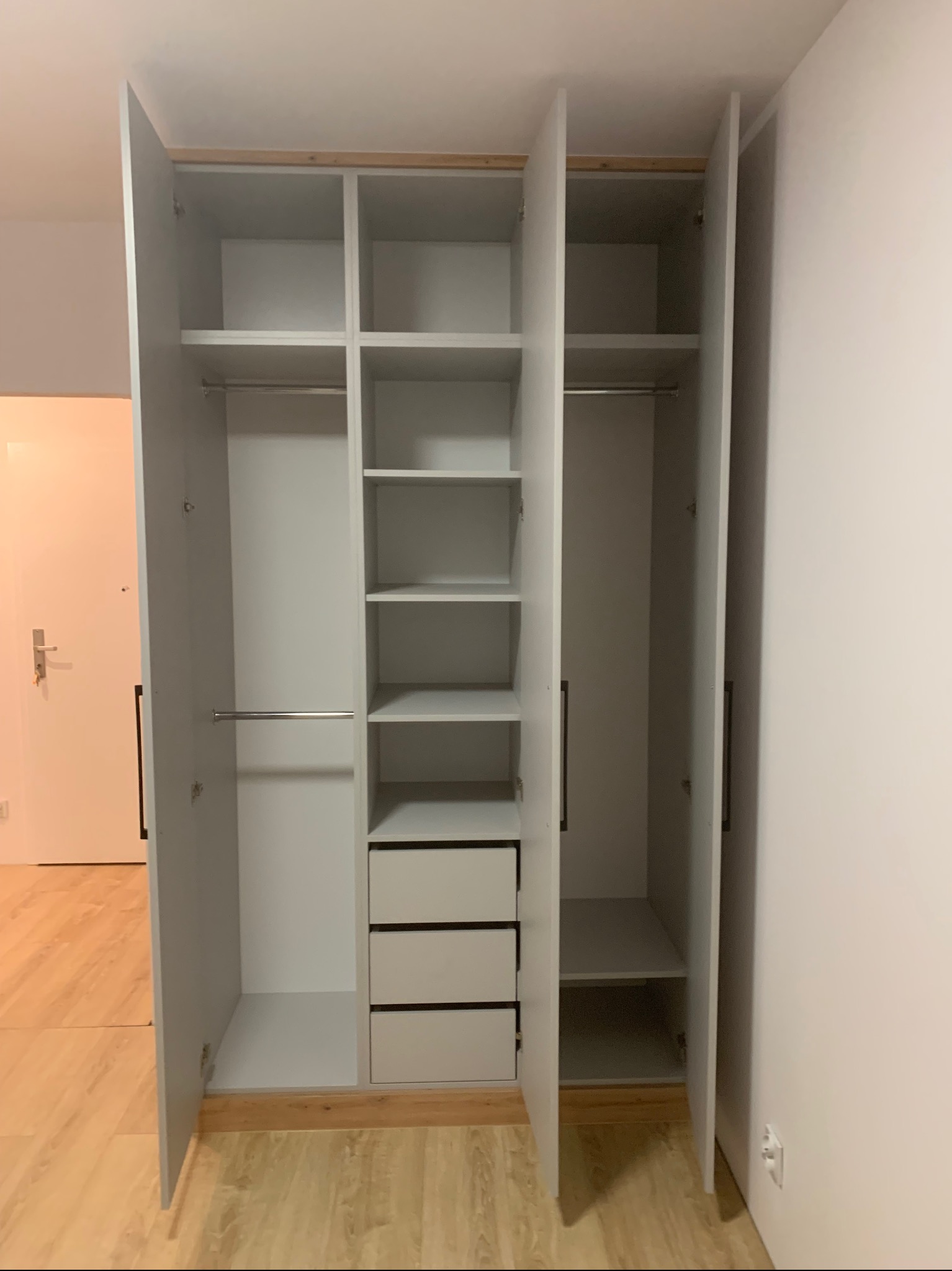 Wnętrze szafy wnękowej z trzema drzwiami, z półkami, drążkami na ubrania i trzema szufladami, w kolorze białym, z widocznym fragmentem podłogi i drzwi wejściowych.