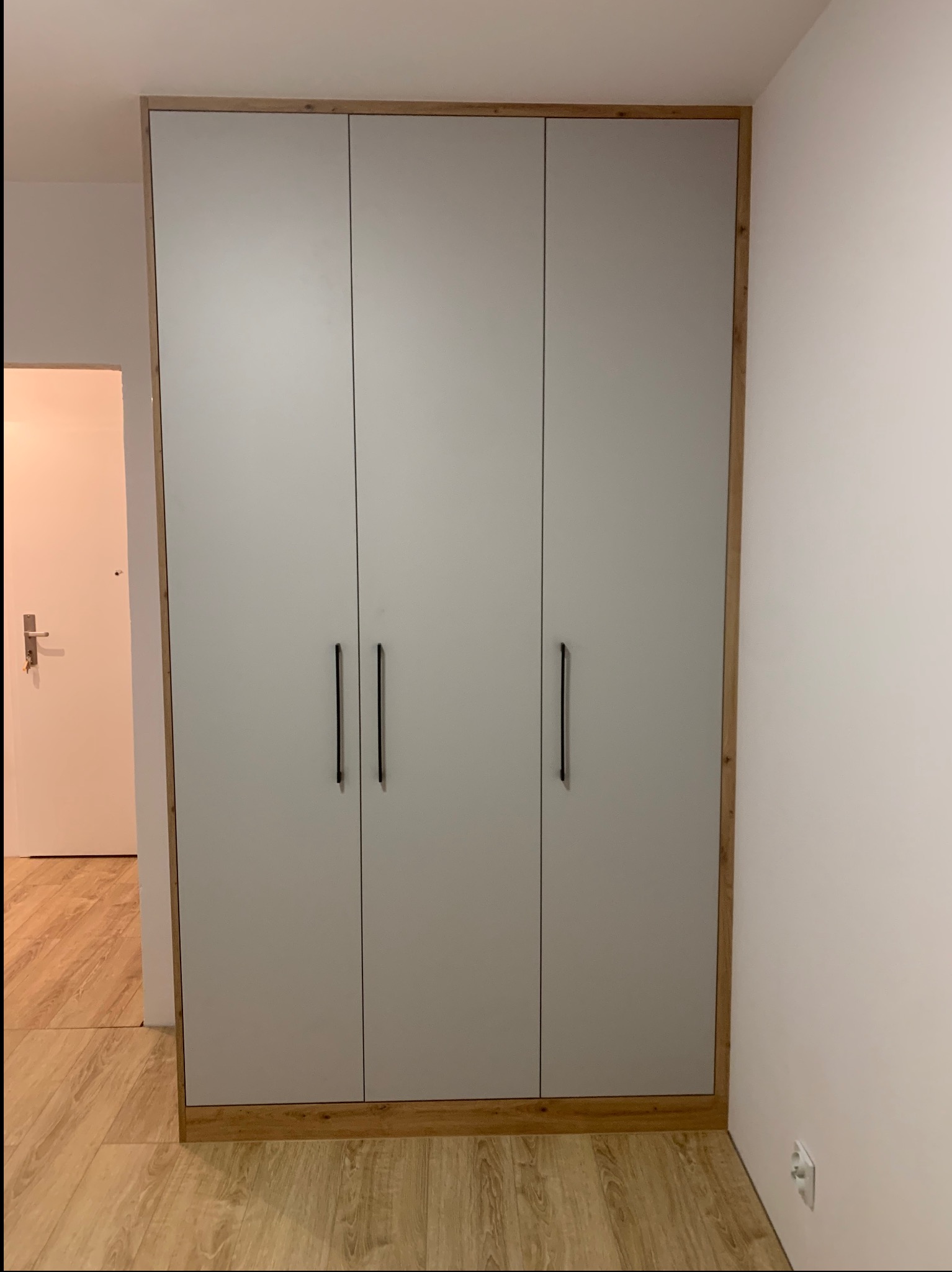 Trzydrzwiowa szafa wnękowa z szarymi frontami i drewnianą ramą, z czarnymi, pionowymi uchwytami, umieszczona w korytarzu z drewnianą podłogą, widoczny fragment białych ścian i drzwi wejściowych.