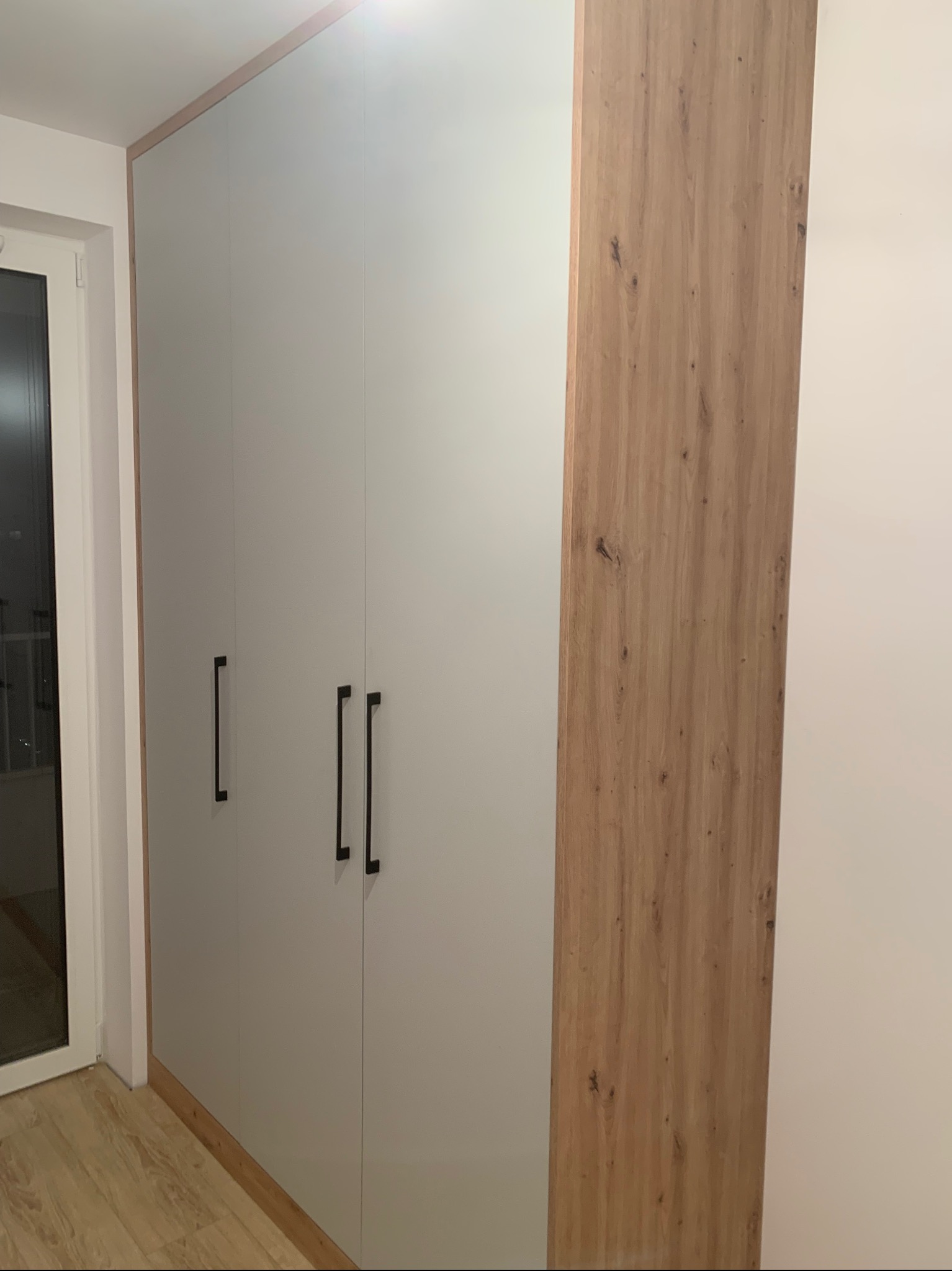 Wysoka szafa z szarymi frontami i drewnianym bokiem, z czarnymi, pionowymi uchwytami, widoczna z boku przy oknie.