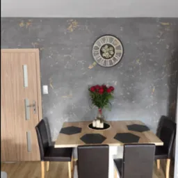 Szara ściana z efektem marmuru i złotymi akcentami, zegar z widocznymi trybikami, bukiet czerwonych r&oacute;ż w wazonie na drewnianym stole z czarnymi podkładkami, krzesła z brązowej sk&oacute;ry, drewniana...