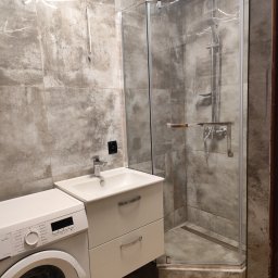 WOJMAR - Nowoczesna łazienka z kabiną prysznicową ze szklanymi drzwiami, umywalką z szafką i pralką, wykończona szarymi płytkami imitującymi beton i drewnopodobną podłogą.