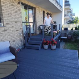 WOJMAR - Niebieski taras z kompozytowych desek, schody i balustrada pomalowane na szaro. Kobieta w białej bluzce i kwiecistej spódnicy stoi na tarasie. Donice z kwiatami.