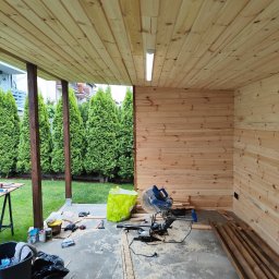 WOJMAR - Wykończenie drewnianej altany: ściany i sufit z boazerii, piła ukośnica, narzędzia, widok na ogród z tujami. Etap prac wykończeniowych.