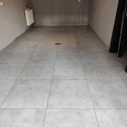 WOJMAR - Wykończona podłoga w szarych płytkach w pomieszczeniu z grzejnikiem i otworem kanalizacyjnym. Widoczne świeżo położone płytki podłogowe w pustym pomieszczeniu.