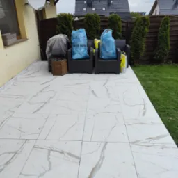Białe płytki imitujące marmur na tarasie, z ciemnymi meblami ogrodowymi i workami na śmieci w tle, widok z góry, jasne światło dzienne.