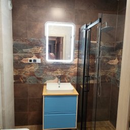 WOJMAR - Nowoczesna łazienka z kabiną prysznicową typu walk-in, umywalką na niebieskiej szafce i podświetlanym lustrem. Brązowe płytki z motywem roślinnym. Widoczny fragment toalety.