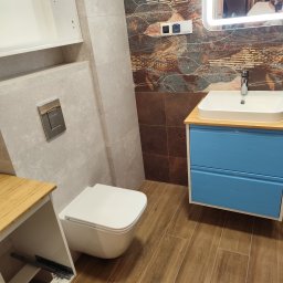 WOJMAR - Nowoczesna łazienka po remoncie: wisząca toaleta, niebieska szafka z umywalką, lustro z podświetleniem LED, brązowe płytki z motywem liści i szare płytki.