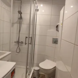 Jasna łazienka po renowacji: kabina prysznicowa ze szklanymi drzwiami, toaleta i białe płytki na ścianach. Widoczne detale wykończenia i armatura.