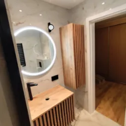 Nowoczesna łazienka po remoncie: drewniana szafka z umywalką, okrągłe lustro z podświetleniem LED i wisząca szafka z lamelami na tle marmurowych płytek.