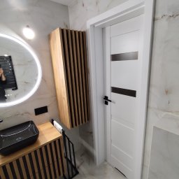 WOJMAR - Nowoczesna łazienka z umywalką nablatową, szafką z drewnianymi lamelami i okrągłym lustrem z podświetleniem LED. Marmurowe płytki na ścianach i podłodze. Białe drzwi z czarną klamką.