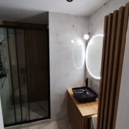 Nowoczesna łazienka z kabiną prysznicową typu walk-in, umywalką nablatową i podświetlanym lustrem. Marmurowe płytki i drewniane akcenty tworzą elegancki wystrój.