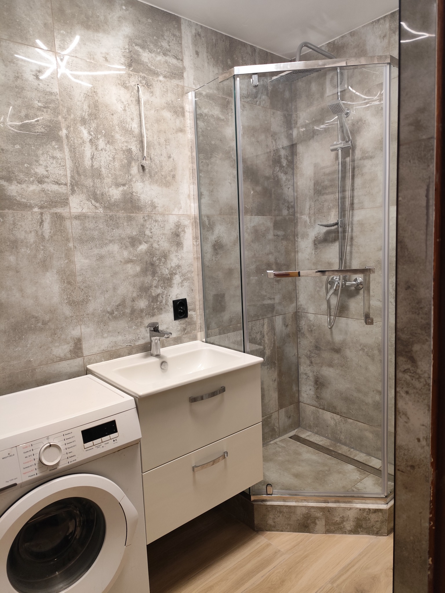 Nowoczesna łazienka z kabiną prysznicową ze szklanymi drzwiami, umywalką z szafką i pralką, wykończona szarymi płytkami imitującymi beton i drewnopodobną podłogą.