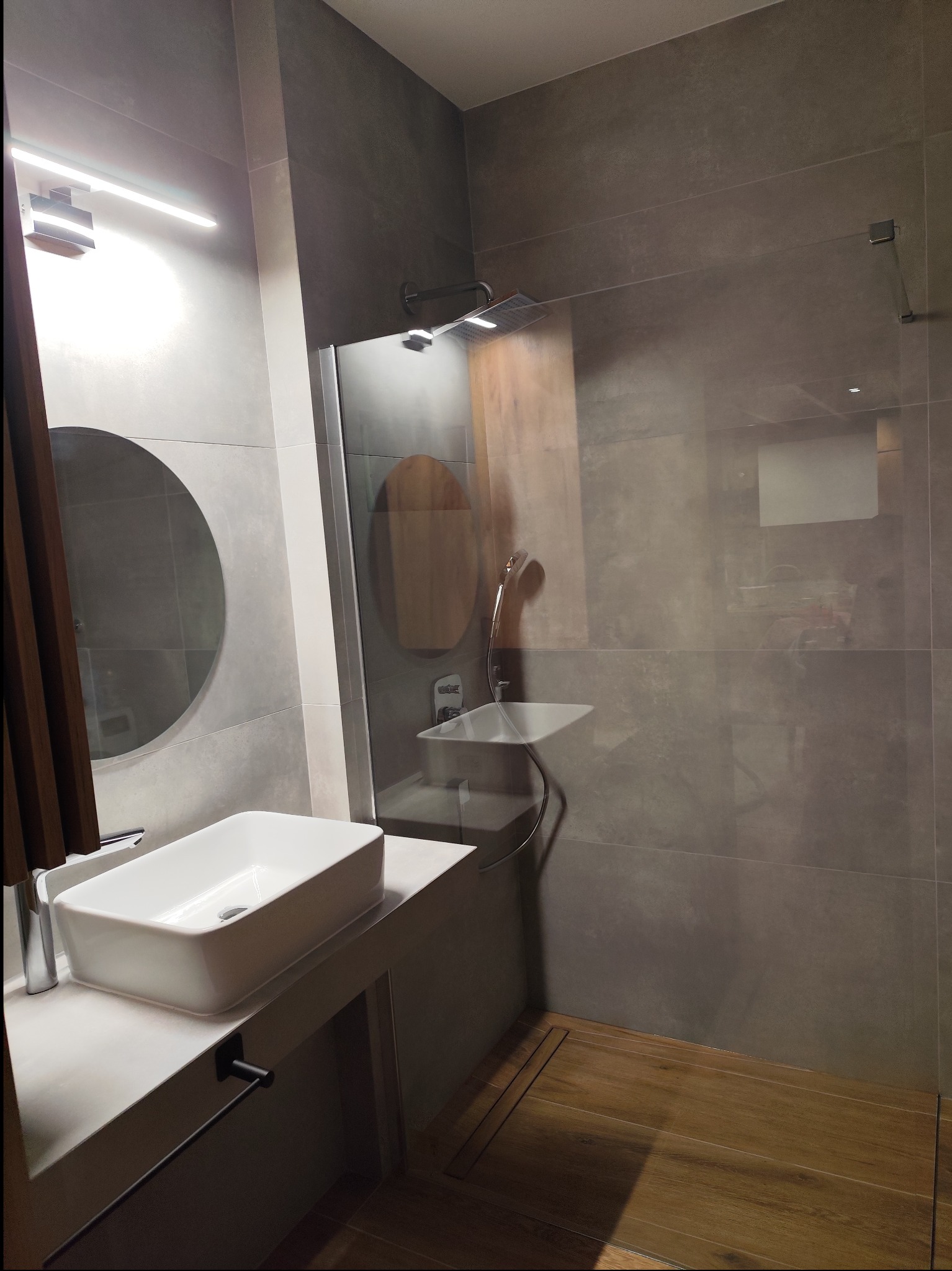 Nowoczesna łazienka z szarą glazurą, drewnianą podłogą w strefie prysznica, okrągłym lustrem i prostokątną umywalką nablatową.