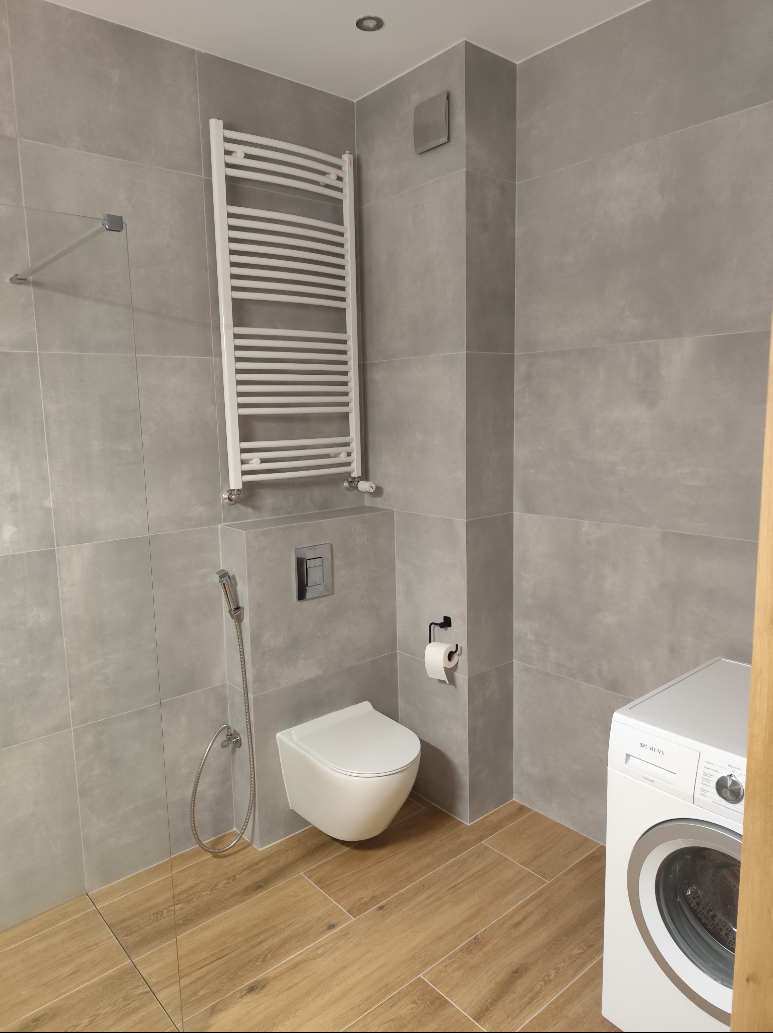 Nowoczesna łazienka z szarymi płytkami, podwieszaną toaletą, grzejnikiem drabinkowym i pralką Siemens, wykończona drewnopodobną podłogą.