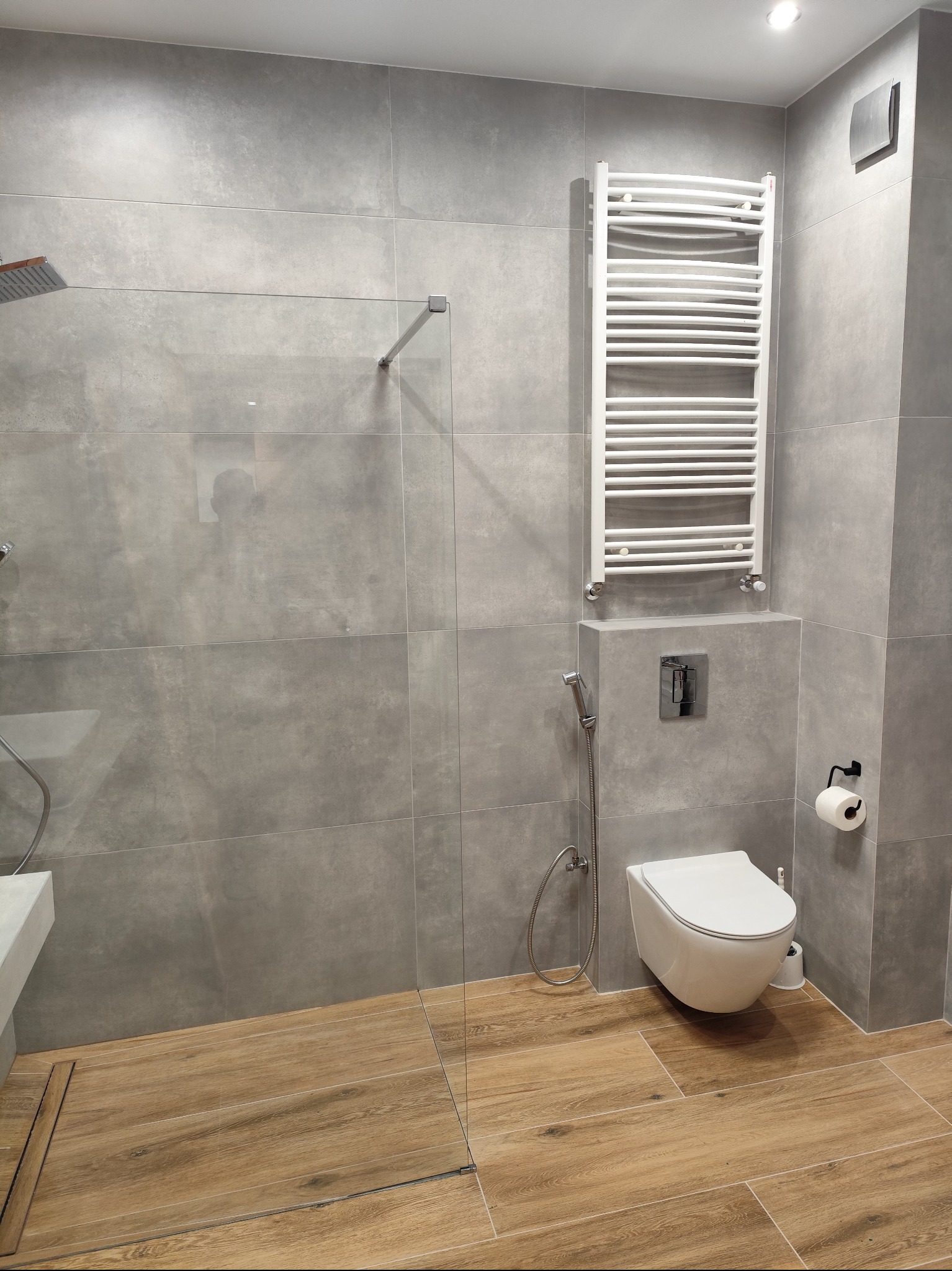 Nowoczesna łazienka z szarymi płytkami wielkoformatowymi, drewnopodobną podłogą, kabiną prysznicową ze szklaną ścianką, podwieszaną toaletą z bidetką i grzejnikiem drabinkowym.