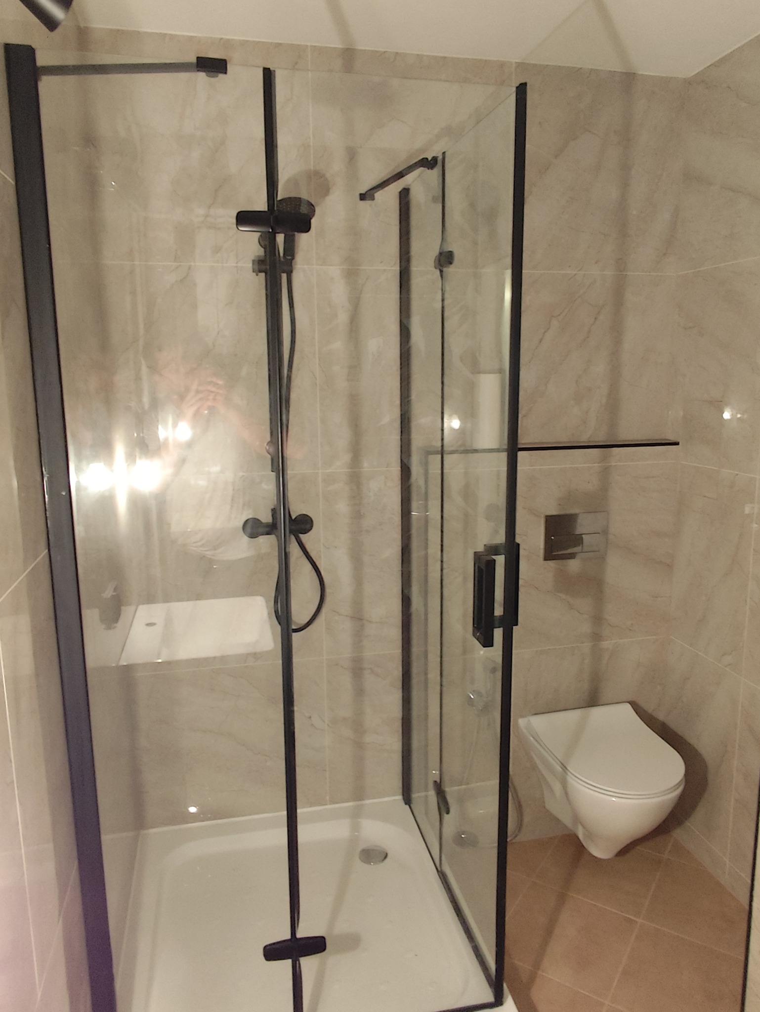 Kabina prysznicowa z czarnymi profilami, armatura w kolorze czarnym, widoczna umywalka i toaleta podwieszana na tle beżowych płytek imitujących marmur.