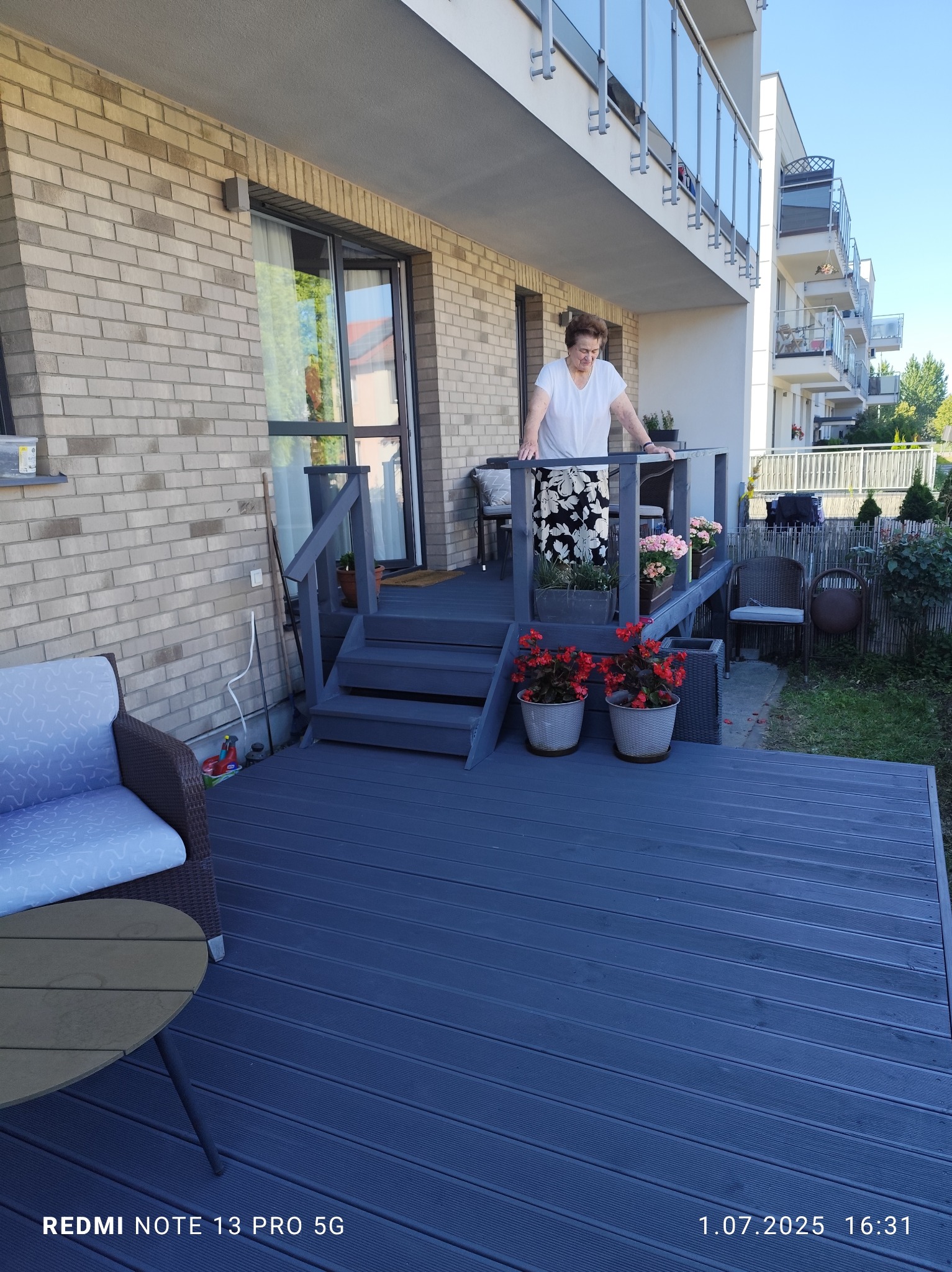 Niebieski taras z kompozytowych desek, schody i balustrada pomalowane na szaro. Kobieta w białej bluzce i kwiecistej spódnicy stoi na tarasie. Donice z kwiatami.