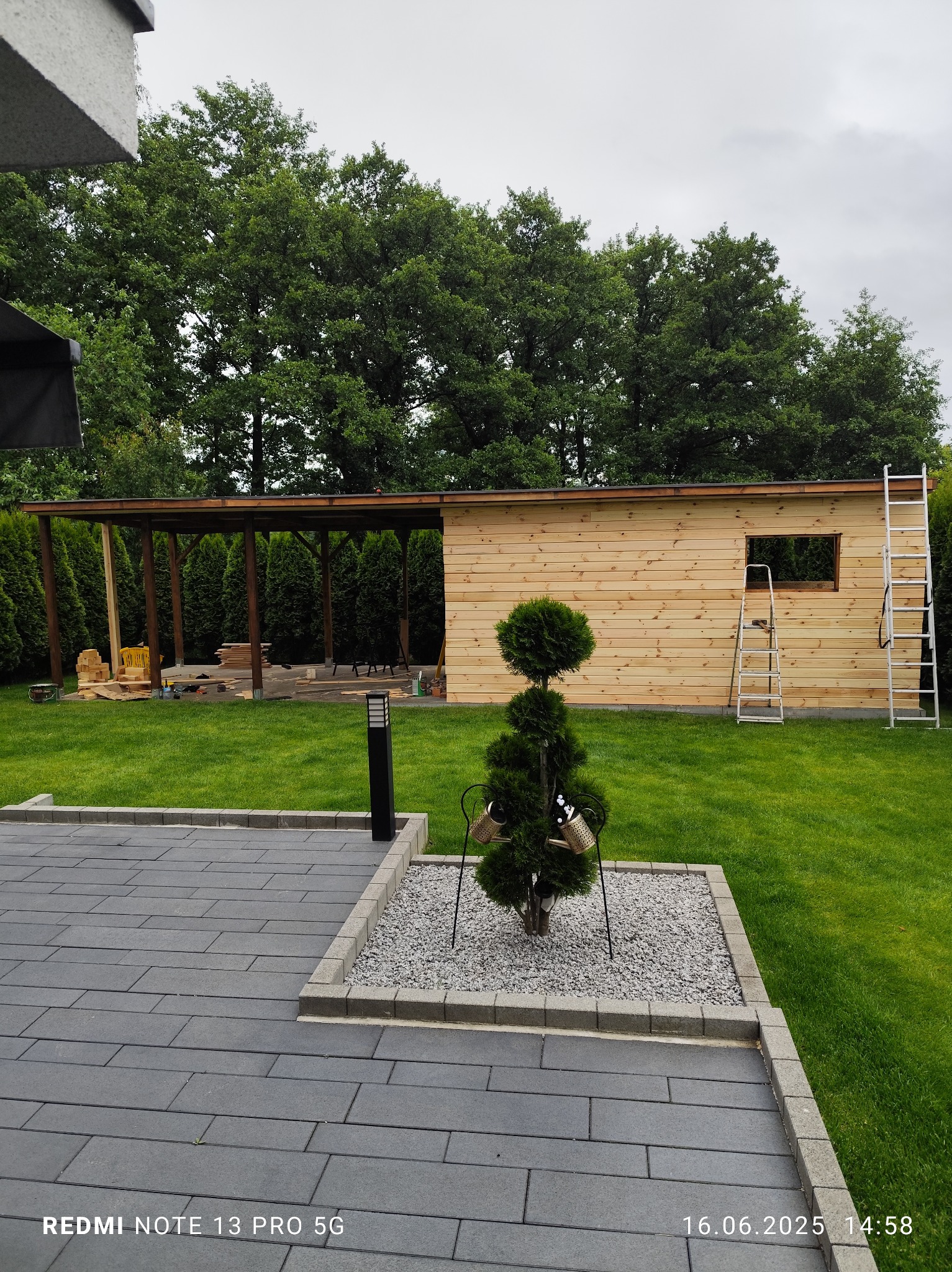 Budowa drewnianego domku narzędziowego w ogrodzie, widoczne drabiny, elementy konstrukcyjne i świeżo ułożona elewacja z desek. W tle zieleń drzew i trawnik.