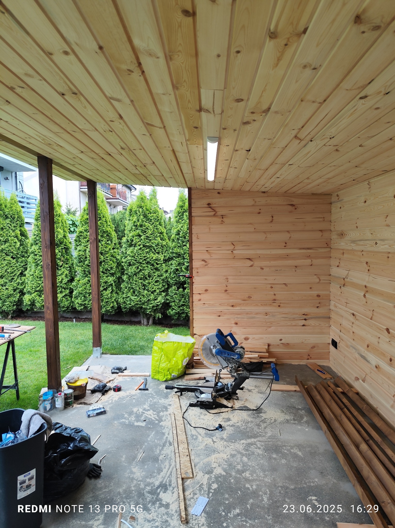 Wykończenie drewnianej altany: ściany i sufit z boazerii, piła ukośnica, narzędzia, widok na ogród z tujami. Etap prac wykończeniowych.