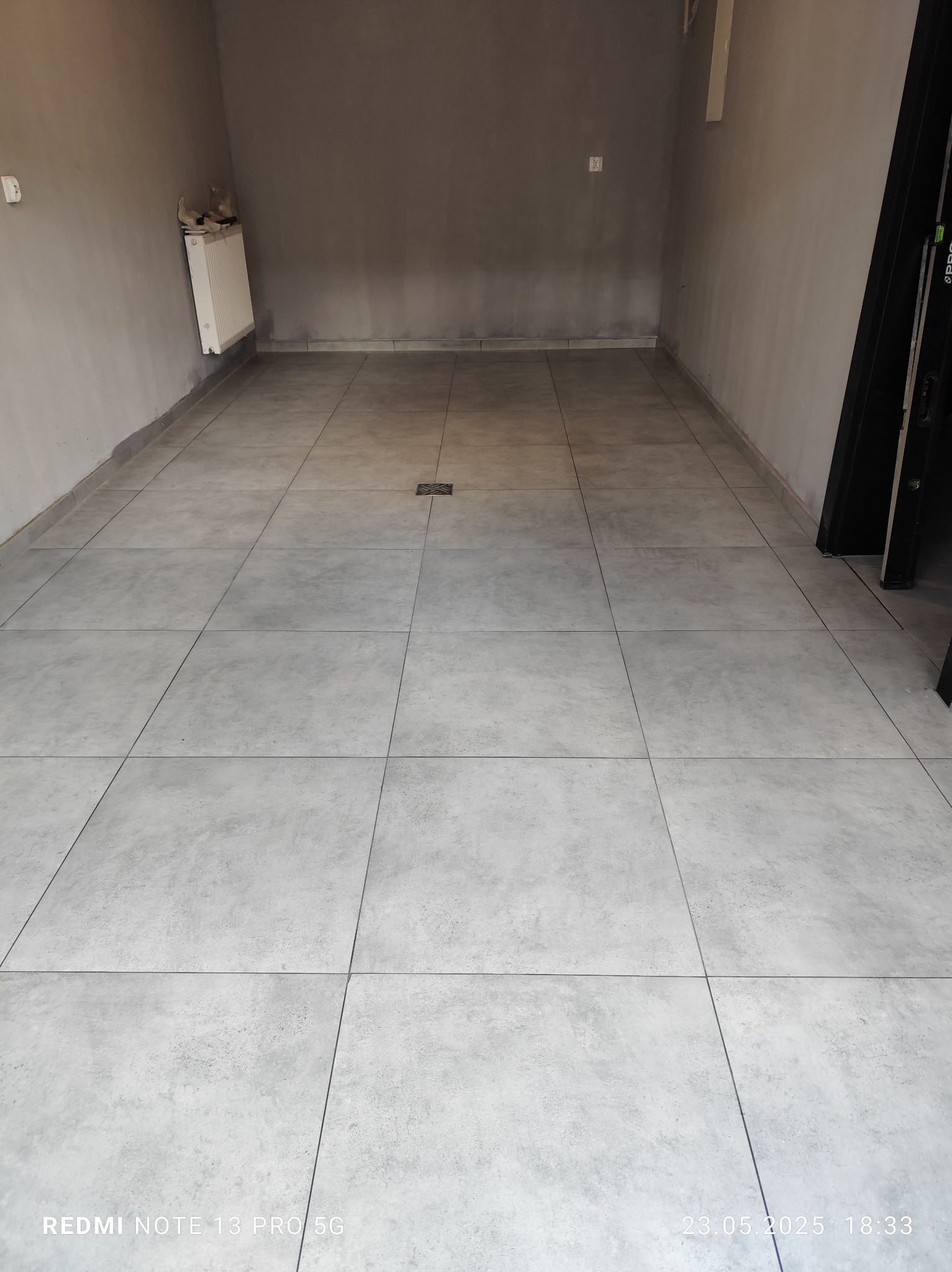 Wykończona podłoga w szarych płytkach w pomieszczeniu z grzejnikiem i otworem kanalizacyjnym. Widoczne świeżo położone płytki podłogowe w pustym pomieszczeniu.