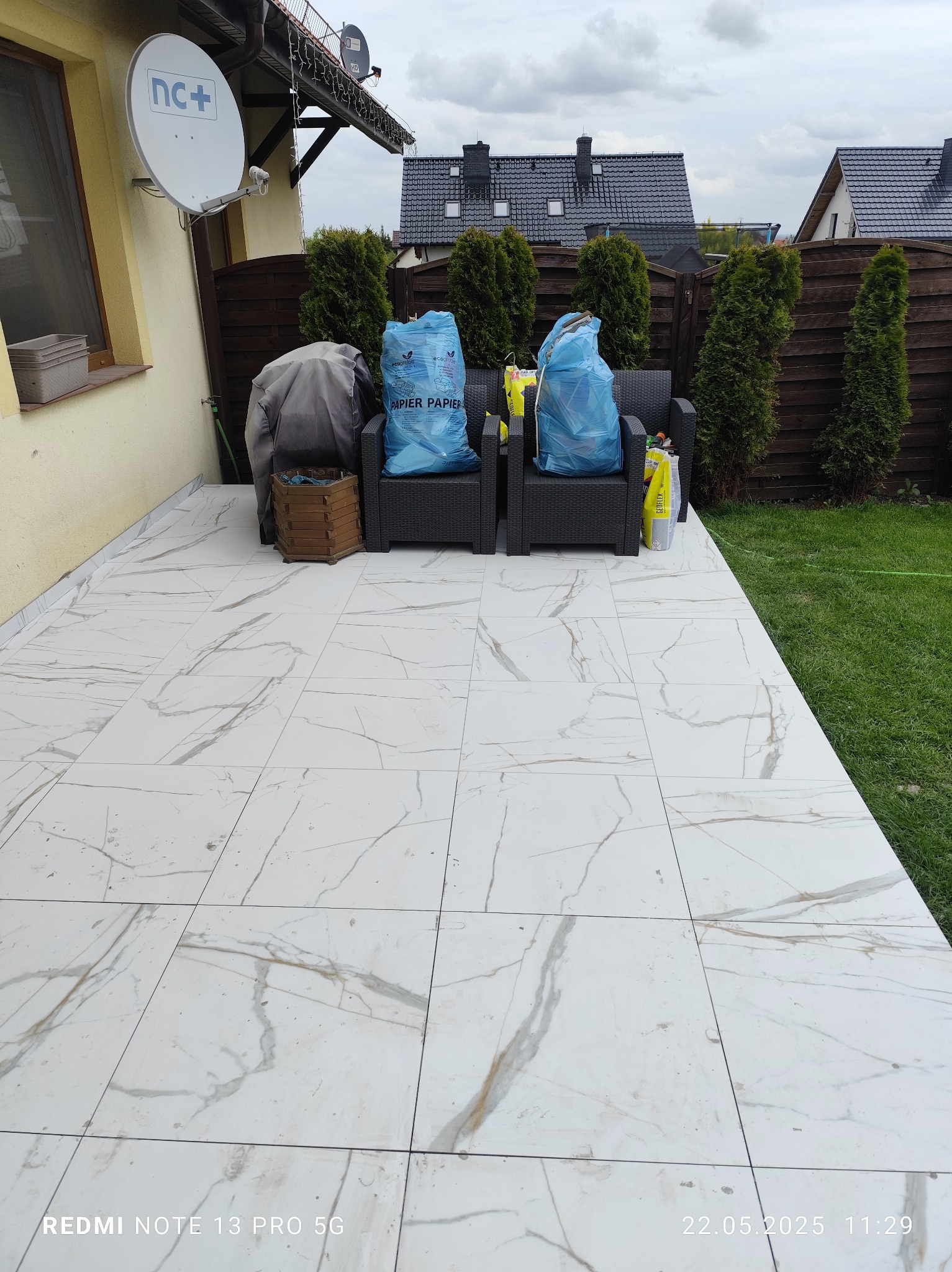 Białe płytki imitujące marmur na tarasie, z ciemnymi meblami ogrodowymi i workami na śmieci w tle, widok z góry, jasne światło dzienne.