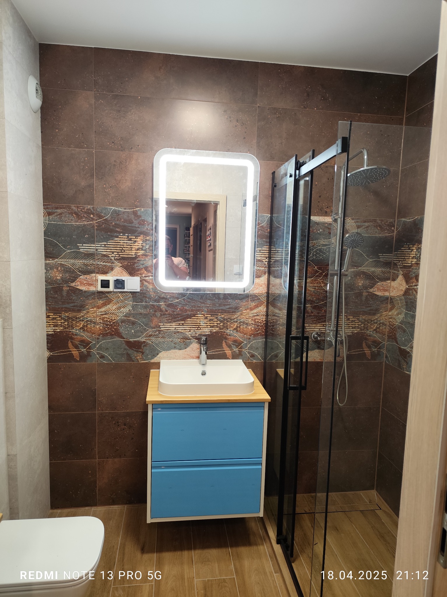 Nowoczesna łazienka z kabiną prysznicową typu walk-in, umywalką na niebieskiej szafce i podświetlanym lustrem. Brązowe płytki z motywem roślinnym. Widoczny fragment toalety.