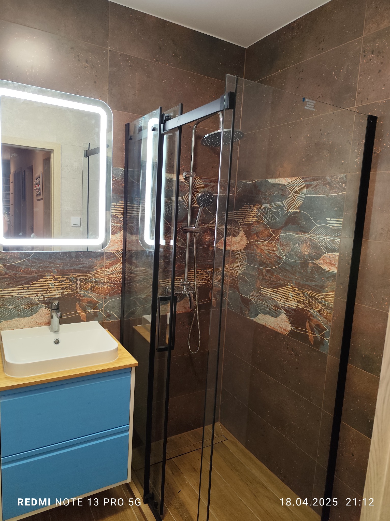 Nowoczesna łazienka z kabiną prysznicową typu walk-in, umywalką na szafce w kolorze błękitu i podświetlanym lustrem. Brązowe płytki z wzorem na ścianach i drewnopodobna podłoga.